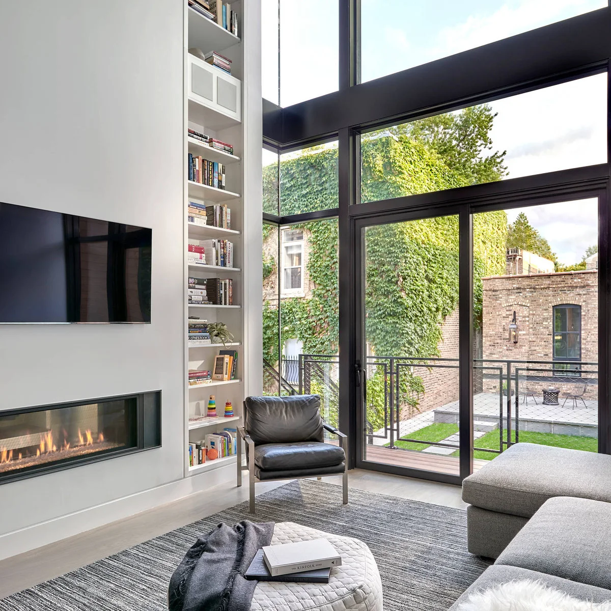 A double-height living room with a narrow full-height bookcase beside tall glazing, a gray sectional and a black chair.