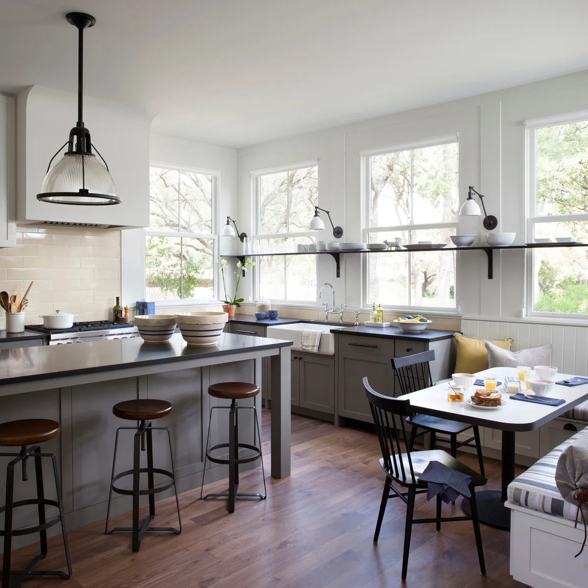 A long dark shelf crosses a row of kitchen windows above a sink, with dishes and small wall lights.