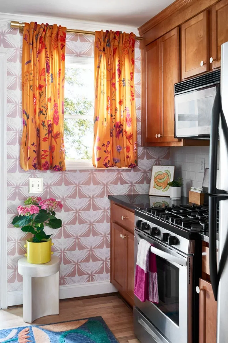 Warm wood cabinets and a stainless range sit beside pink patterned wallpaper and bright orange floral curtains.