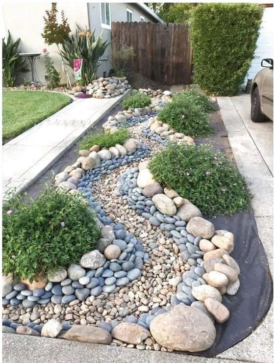 Mixed rounded rocks and low shrubs fill a narrow curved bed between a driveway and entrance path