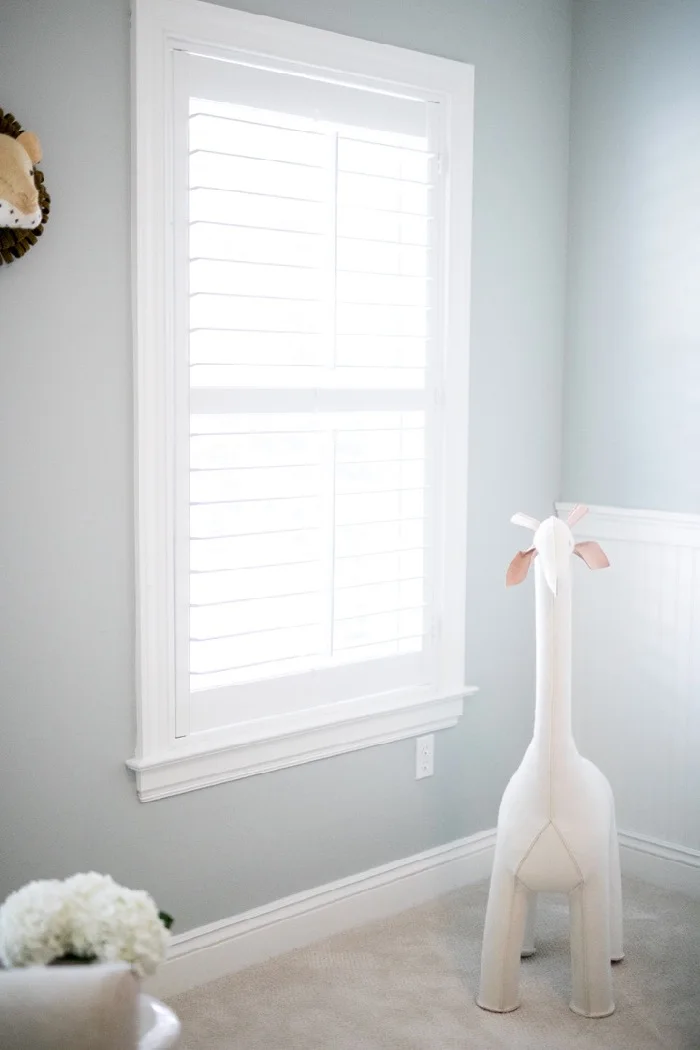 A pale fabric giraffe stands beside a shuttered window against a soft gray nursery wall.