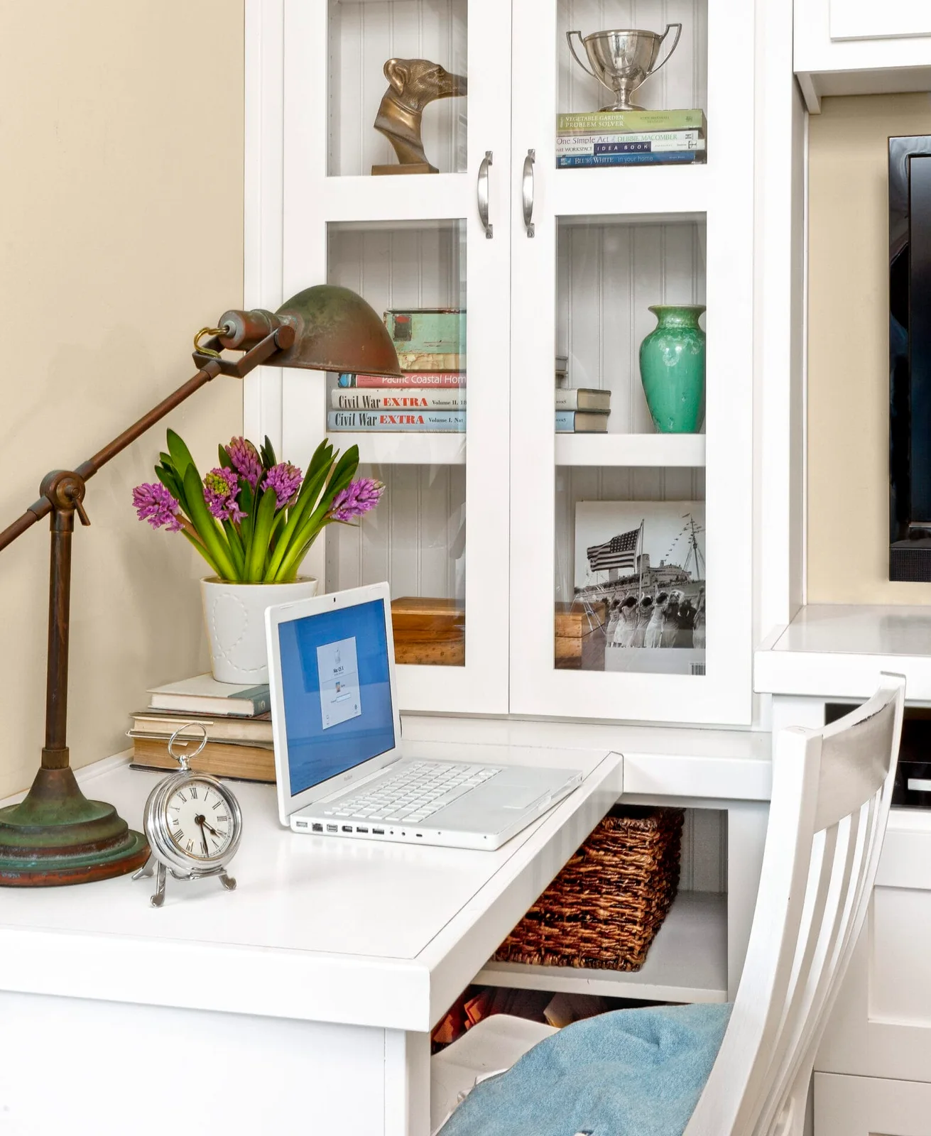 A white built-in desk holds a laptop, a small clock and a desk lamp beside glass-front cupboards in a shared guest room.