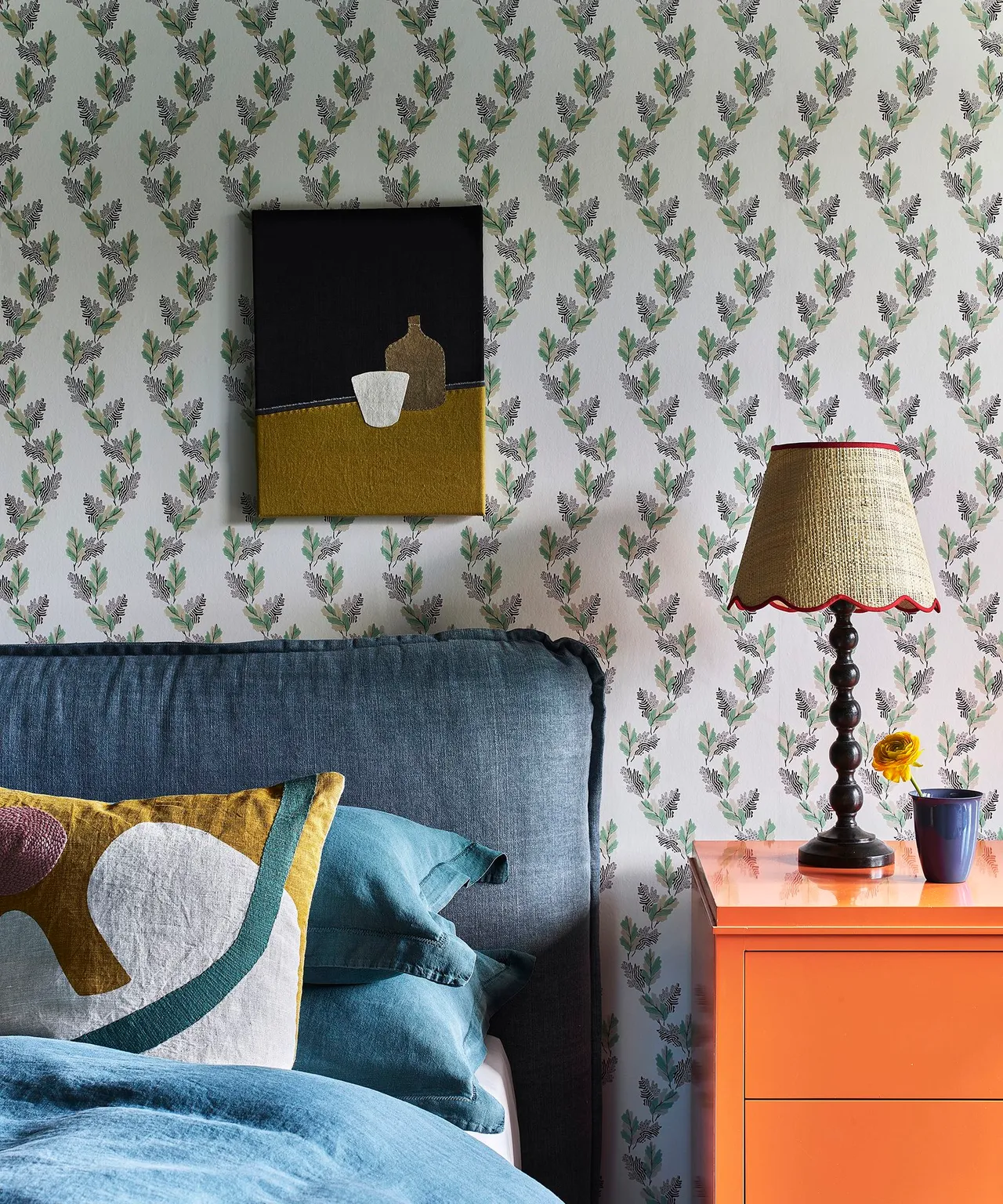 Bedroom with small botanical wallpaper, dark blue headboard and bedding, orange bedside cabinet, turned dark lamp and framed abstract picture.