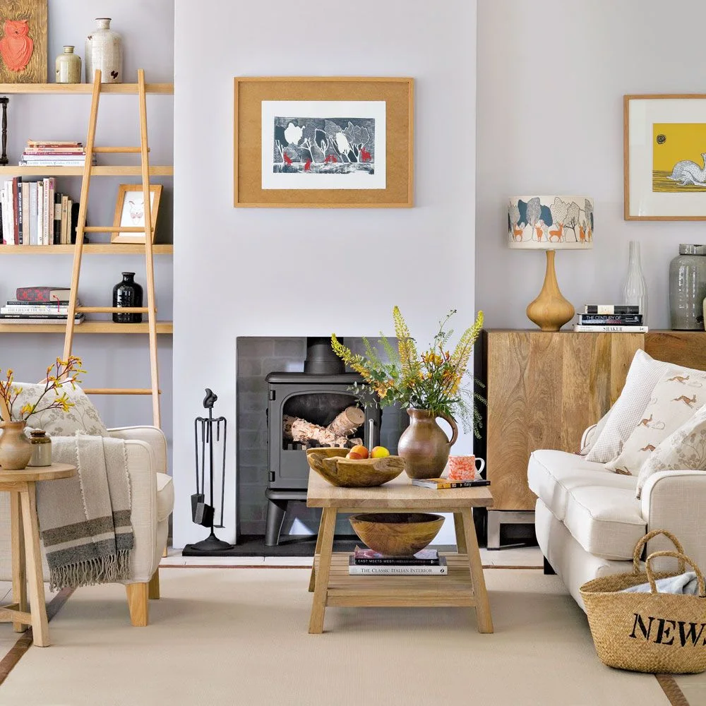 Light wood shelves and a cabinet flank a dark stove beside pale chairs, a wood coffee table and a woven basket.
