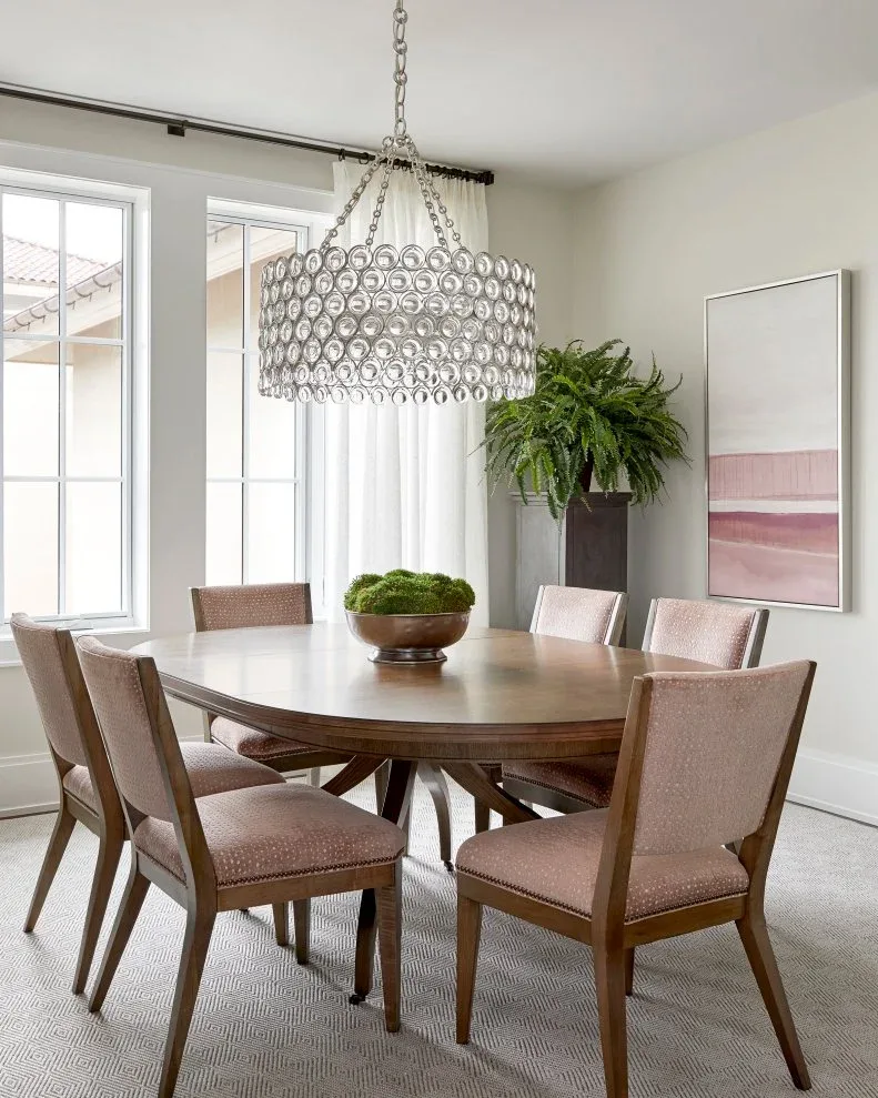 Light dining room with a round wood table, dusty pink chairs, a circular-loop drum chandelier and pale pink abstract artwork