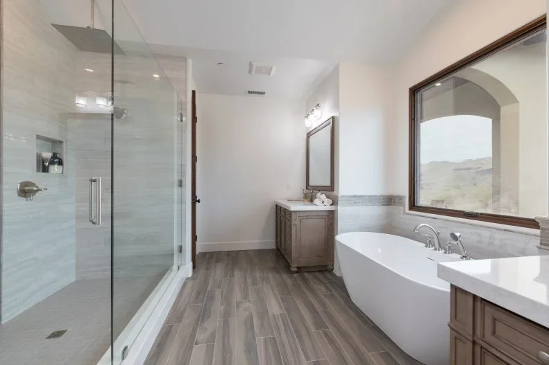 A pale bathroom has a glazed shower on one side, a white tub below a large framed picture and wood-colored vanity storage along the other wall
