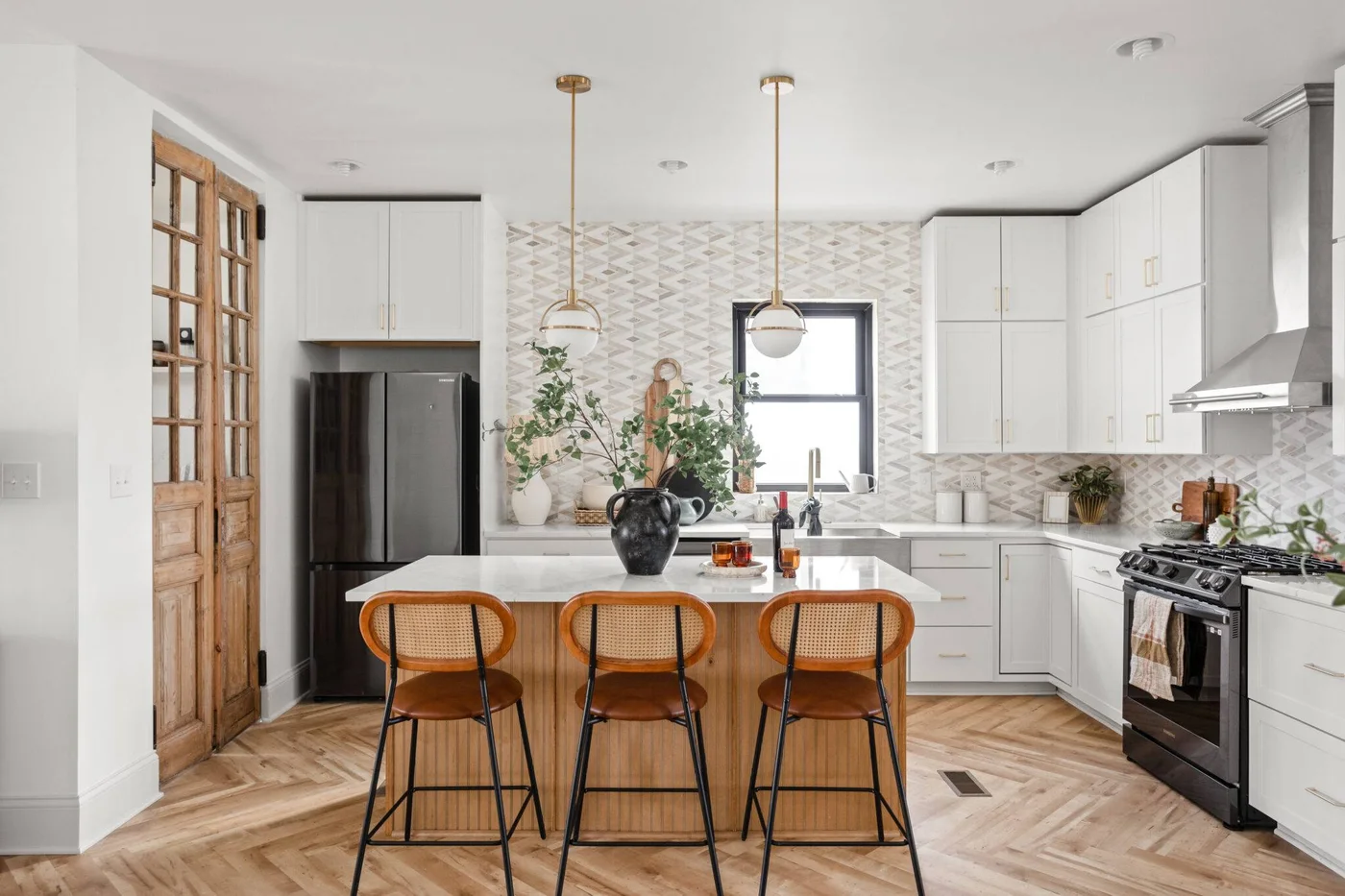 A white kitchen has a pale diamond-patterned mosaic backsplash beside timber doors, a timber island front and woven stools.
