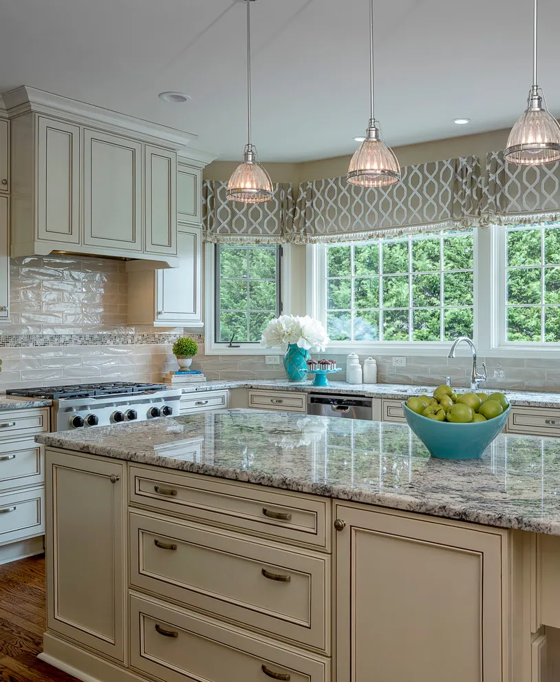 Cream-beige cabinets with glass pendants and a patterned Roman shade above the sink