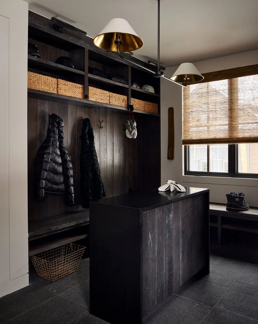 Dark wood mudroom lockers with coat hooks, upper woven baskets, a central bench unit, side seating, gray floor, window shade, and metal pendant lights.