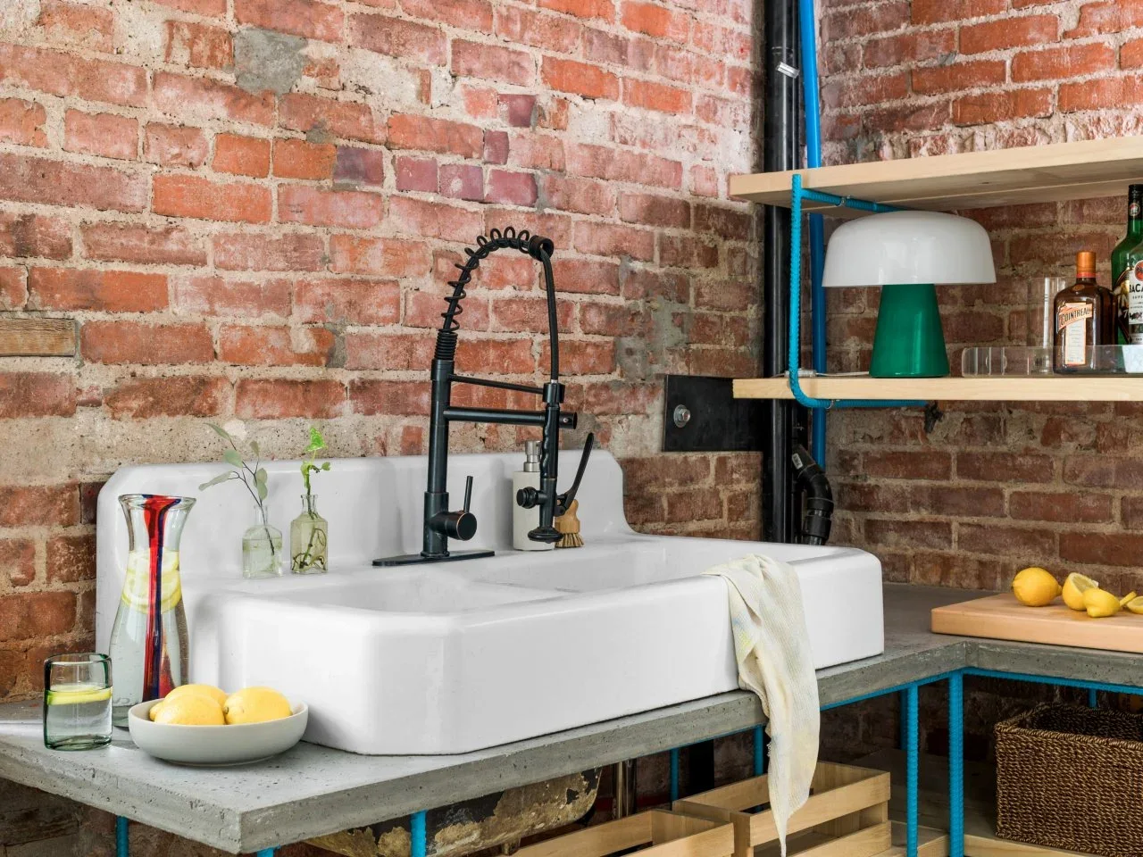 A white apron-front sink stands against exposed brick with timber shelves, blue supports and storage crates below.