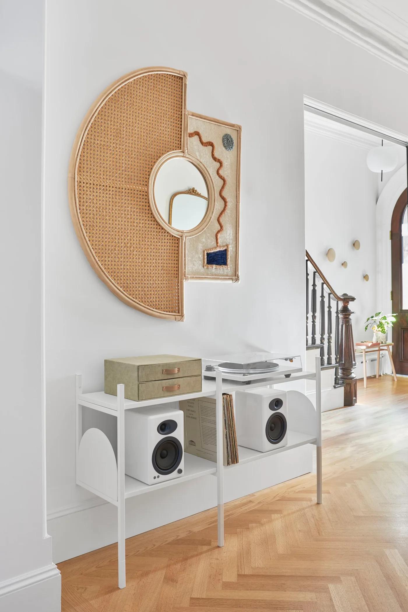 Pale living room audio corner with woven semicircular wall piece, small round mirror and white equipment stand