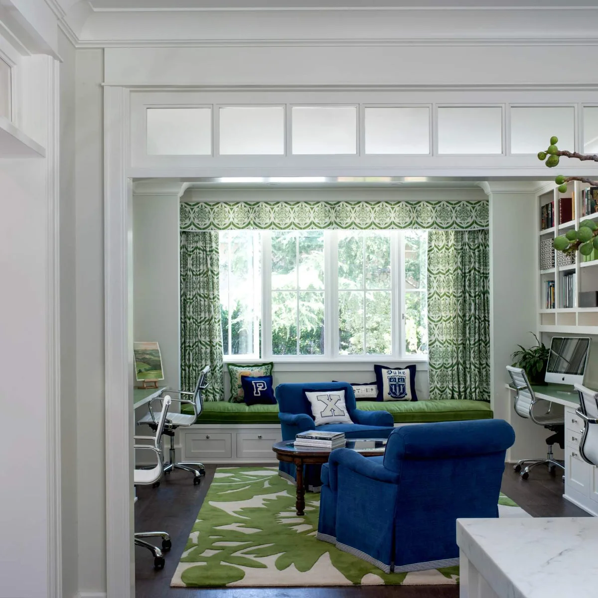 Shared office with desks along the sides, two blue armchairs, green patterned rug and long green window seat