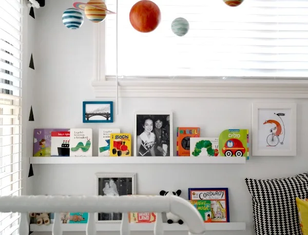 Pale nursery book ledges hold colorful covers and framed pictures beside a patterned dark pillow and pale window treatment