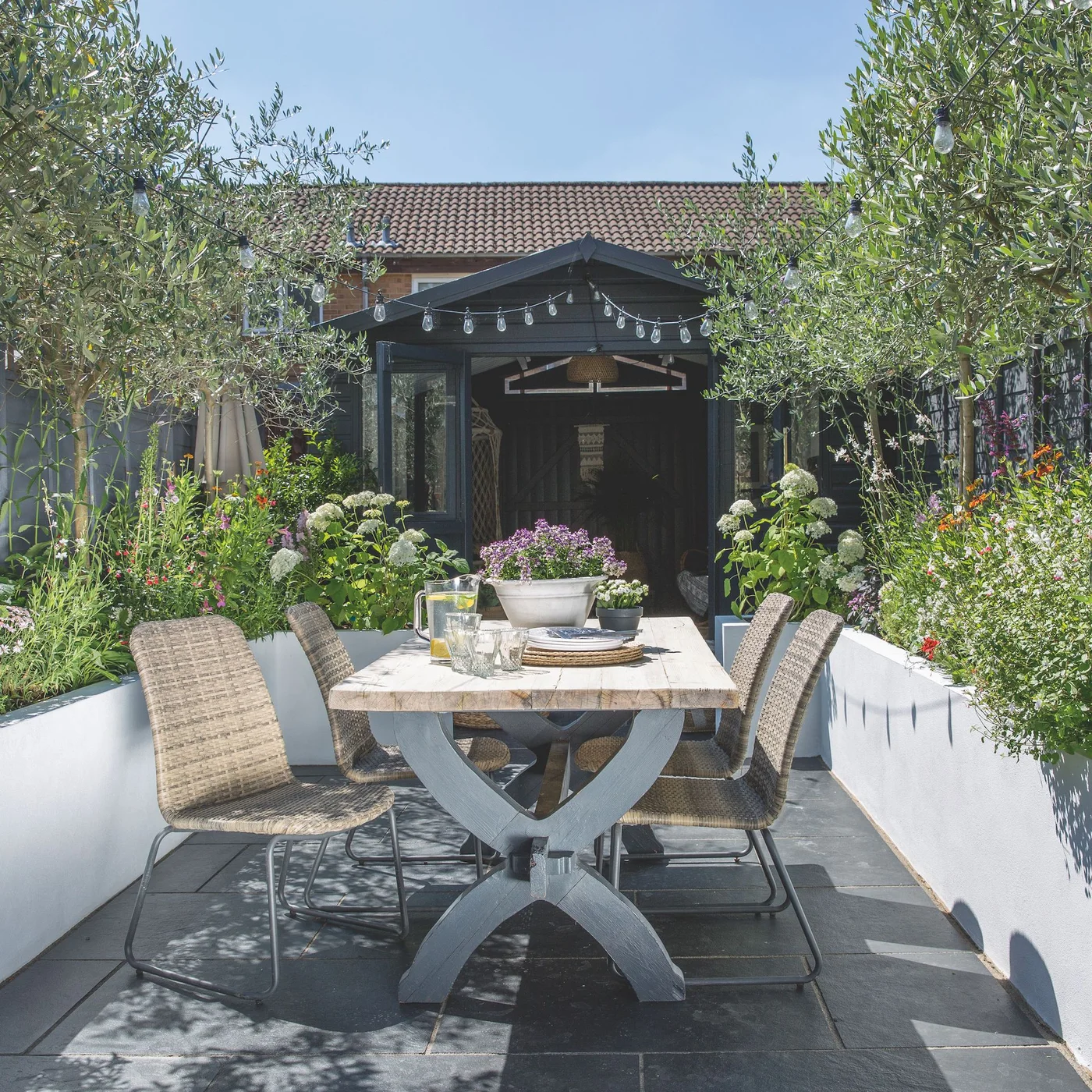 A dining table stands between pale raised beds with flowering plants and slender trees