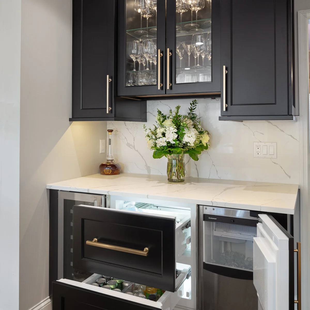 Black mini bar with open refrigerated drawers, open ice-machine door, pale counter and glass-front upper cupboards
