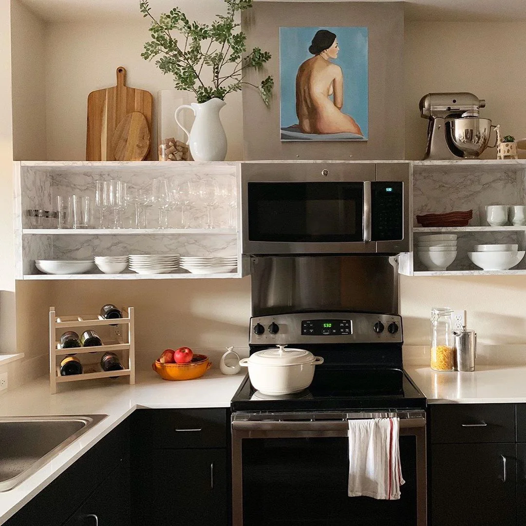Boards, a white pitcher, a small jar and a figure painting above open kitchen cabinet boxes with a mixer at the far right.