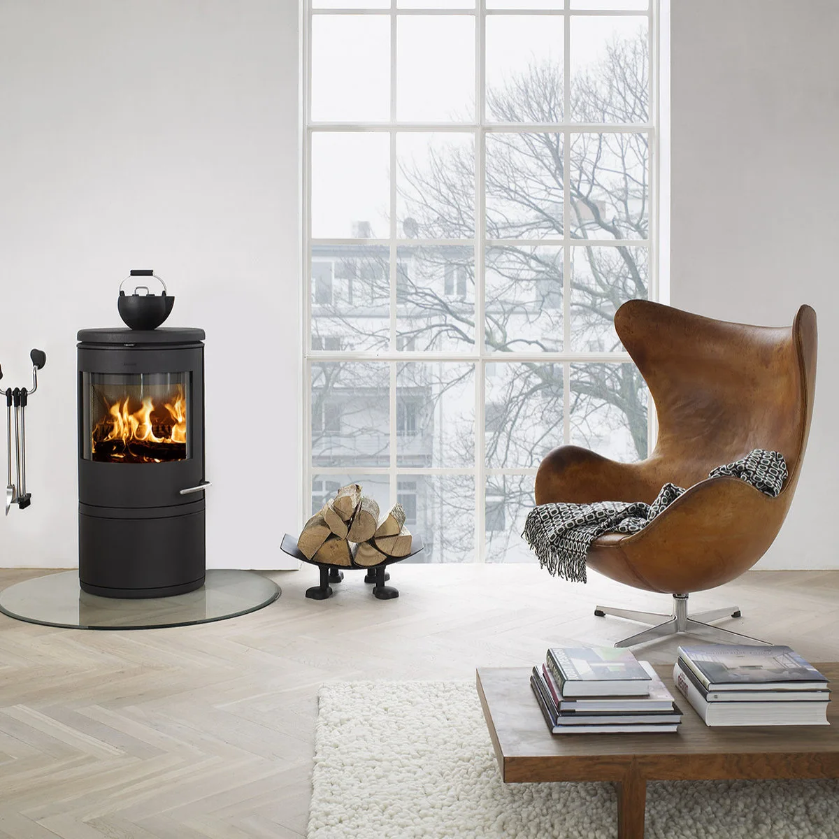 A cylindrical black stove on a round transparent floor panel sits beside a brown wingback chair in a pale room with a tall window.
