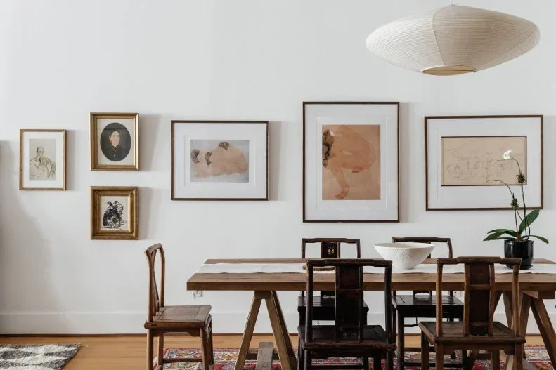 Mixed framed figure drawings and portraits on a white wall above a rustic wooden table, with mismatched wooden chairs, an oval paper pendant, and an orchid.