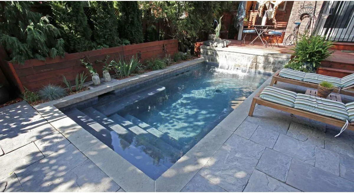 A compact rectangular pool has walk-in steps, underwater benches, a raised waterfall wall and two loungers on the adjacent paving.