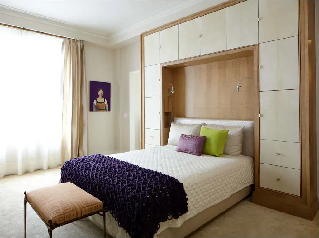 Bed surrounded by pale fitted cupboards and drawers within a wood-framed headboard wall