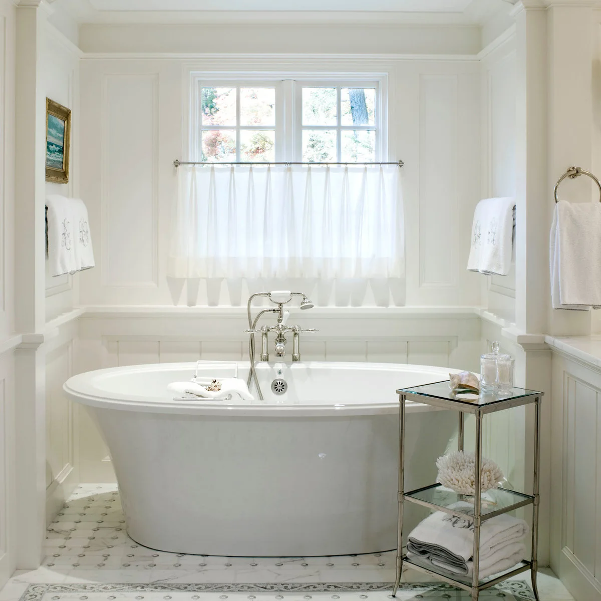 A short white cafe curtain spans a window above a freestanding white bath with white wall paneling.
