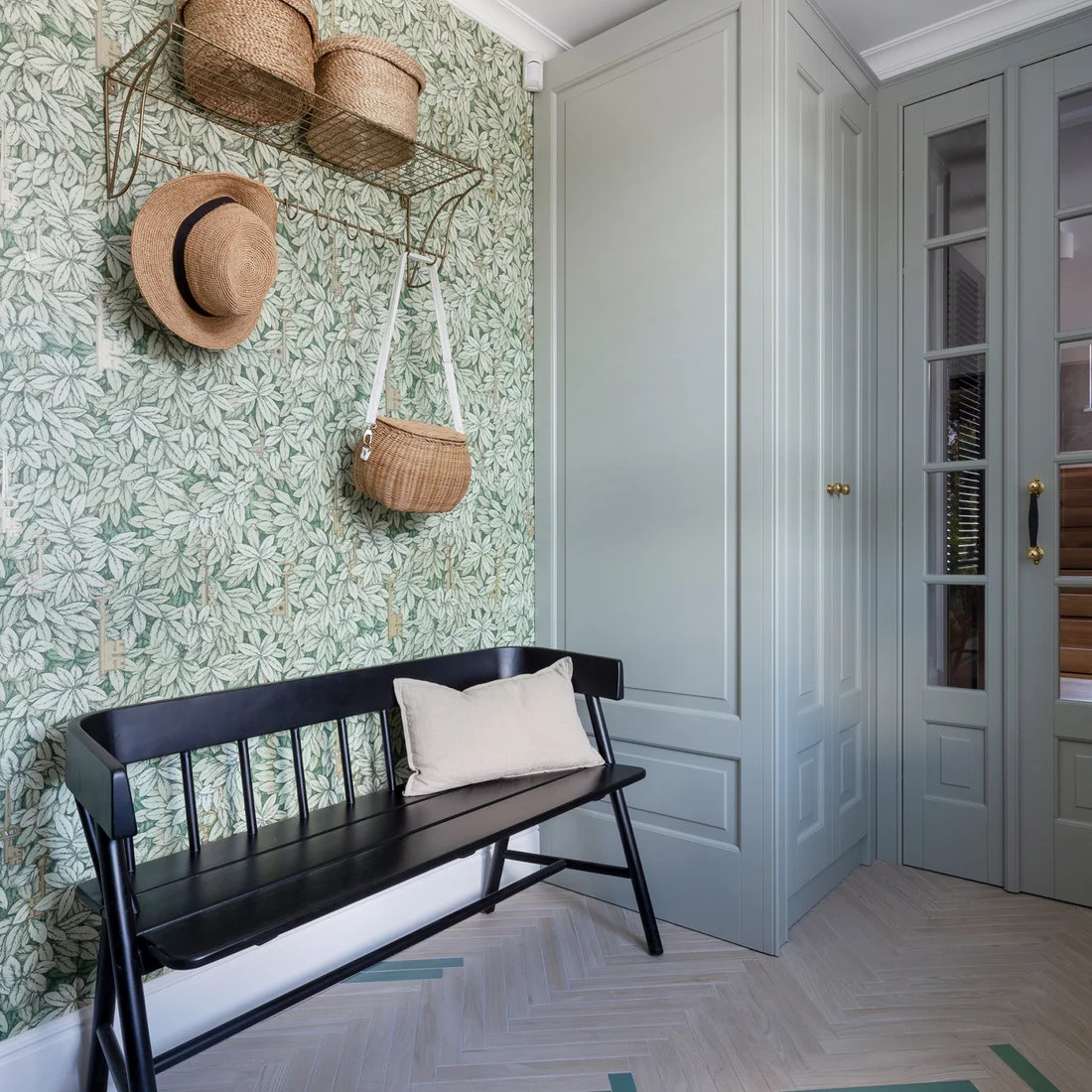 A black spindle-back bench stands against green-and-white leaf wallpaper beside gray cupboards with a woven hook shelf above.