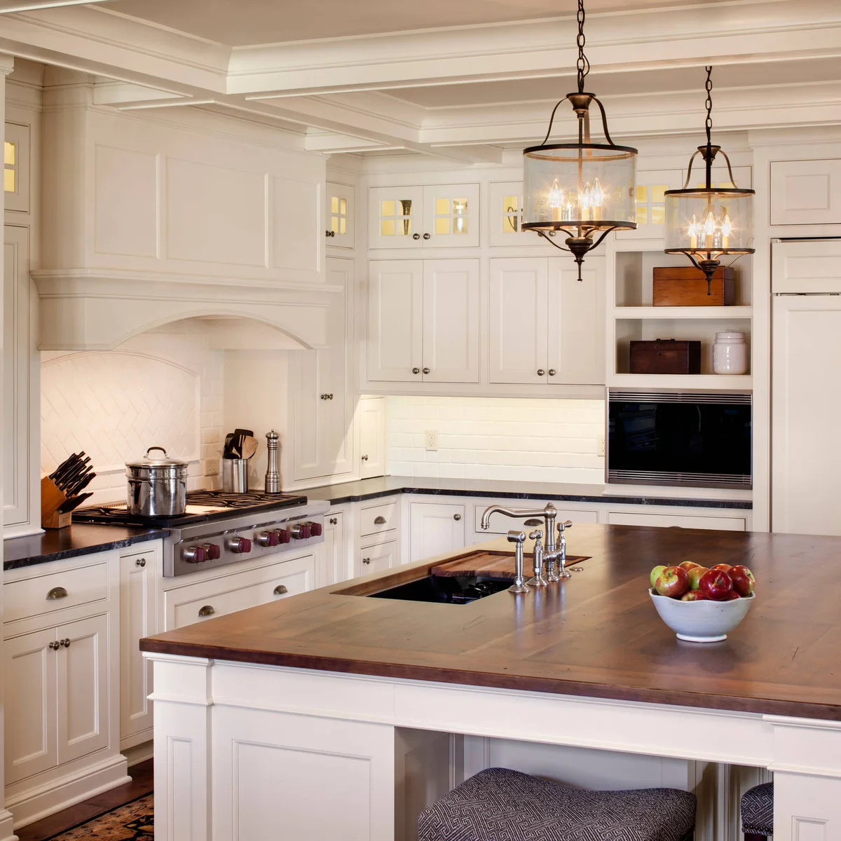 Two dark-framed cylindrical glass lanterns hang above a wood-topped island beneath a pale coffered kitchen ceiling.