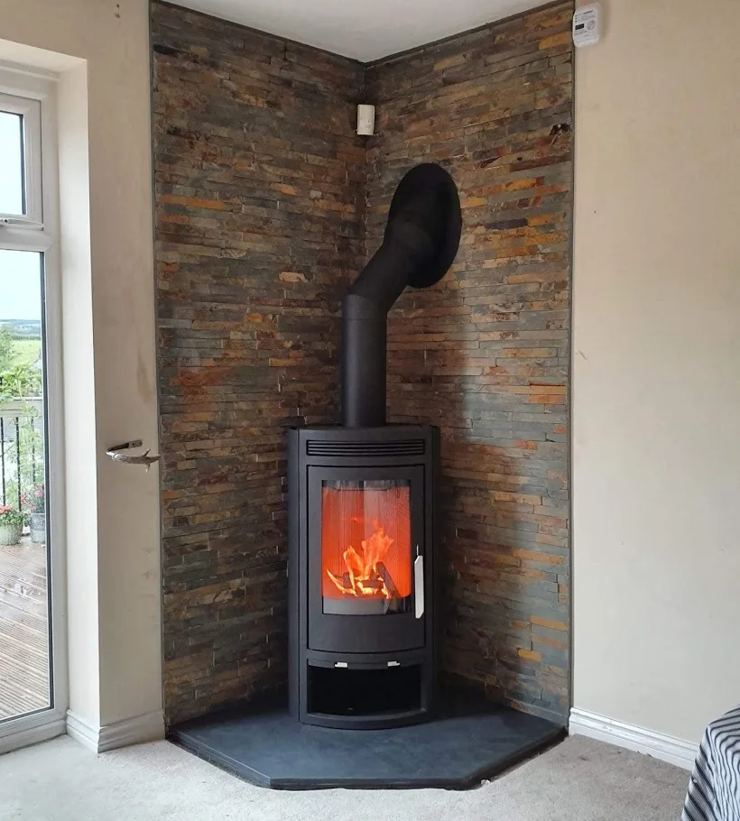 A cylindrical black stove stands on a broad five-sided slate hearth against a corner lined with varied stone slips.