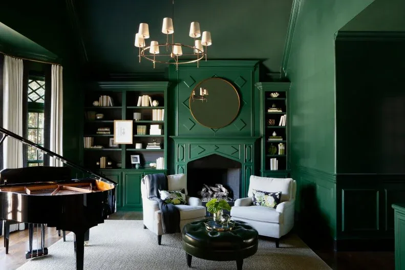Deep green fitted bookcases around matching fireplace and round mirror with pale chairs and black piano