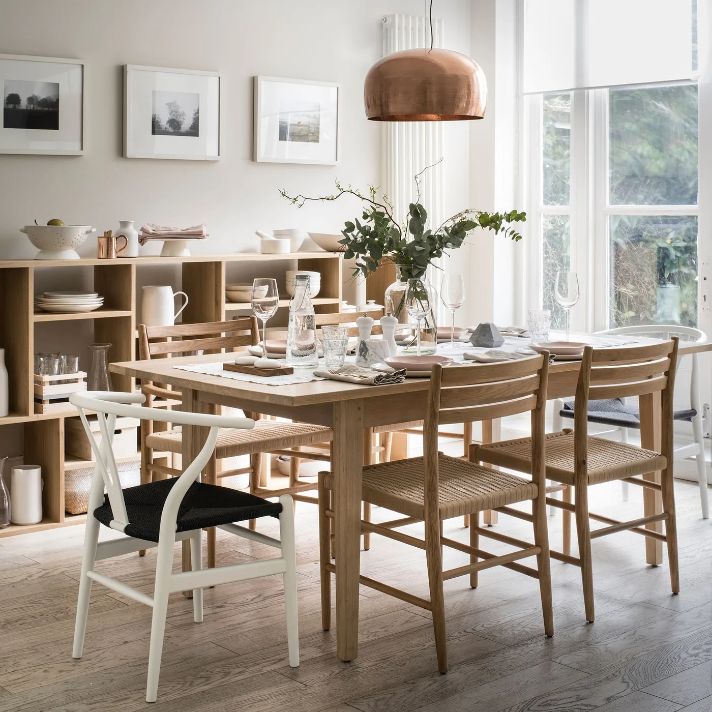 Country dining room with wooden table and chairs, low open cubbies holding dishes, pale walls, framed pictures and metallic pendant shade.