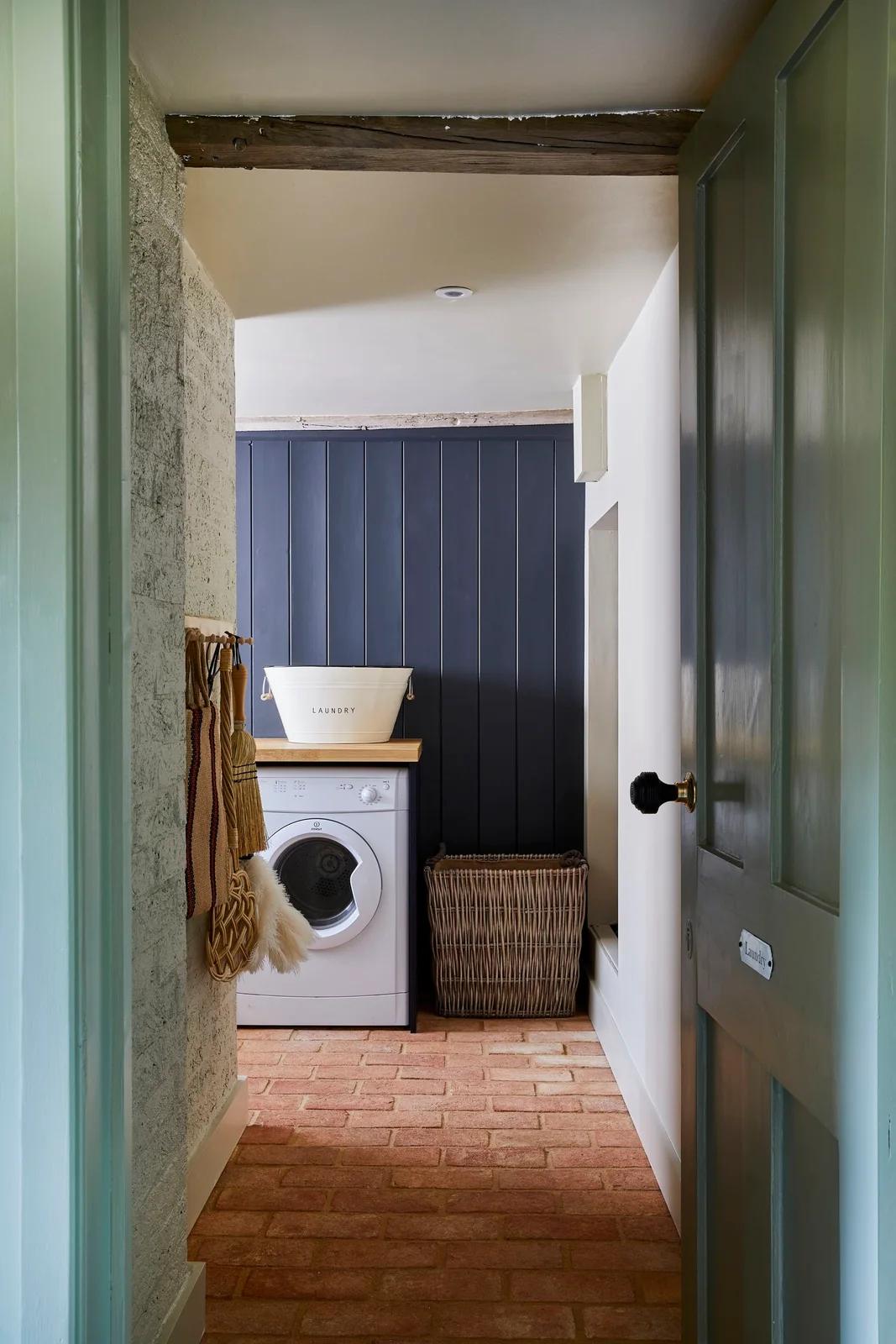 A green doorway opens to a small laundry nook with a white washer beneath a wood counter, dark blue paneling, wall hooks and a woven hamper.