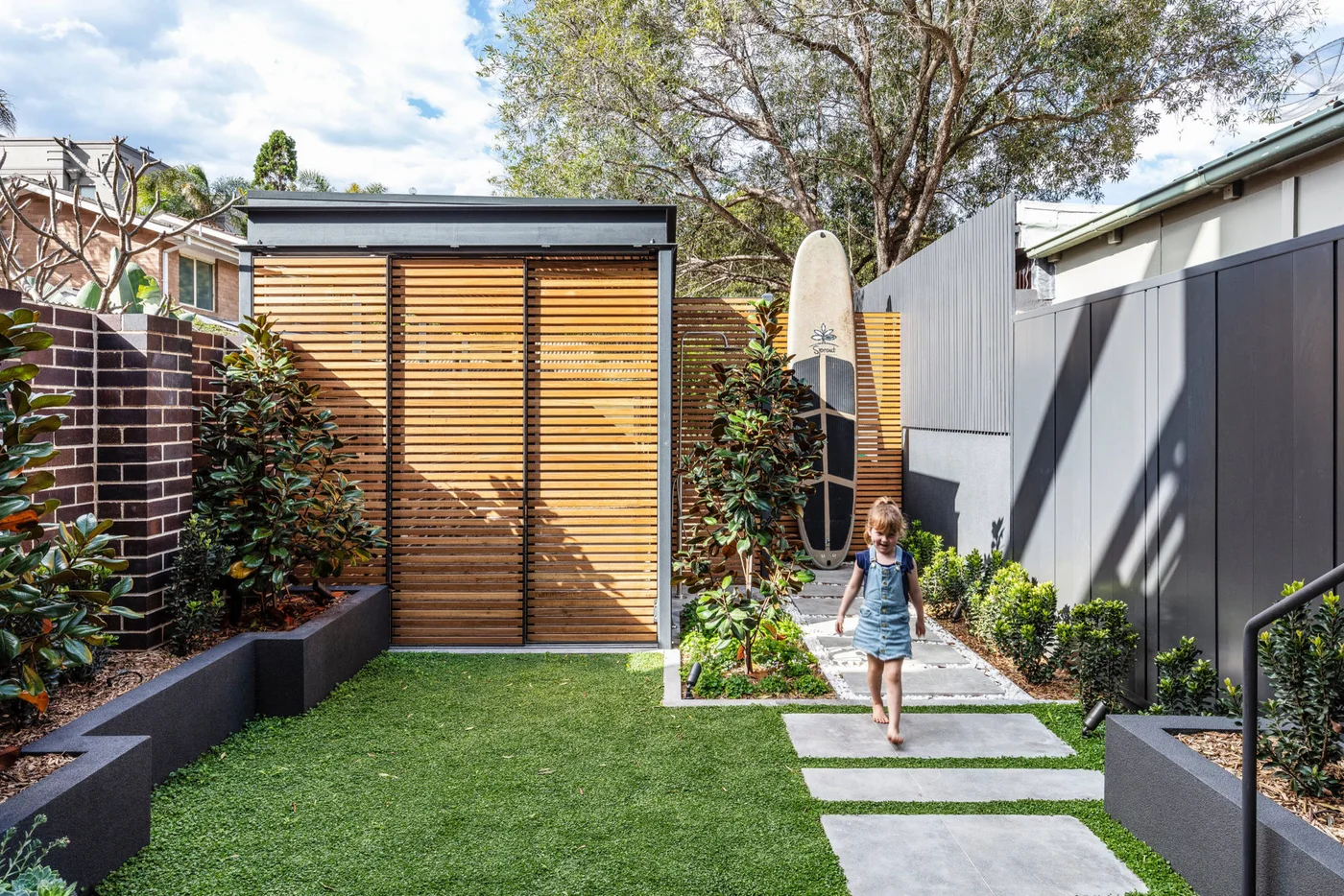Large rectangular slabs run beside a small lawn toward a timber-screened garden structure