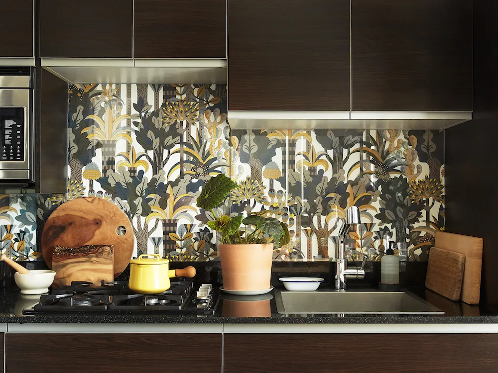 Dark brown flat-front cabinets above and below a black counter, botanical patterned backsplash, metal sink, yellow kettle, and wooden boards.