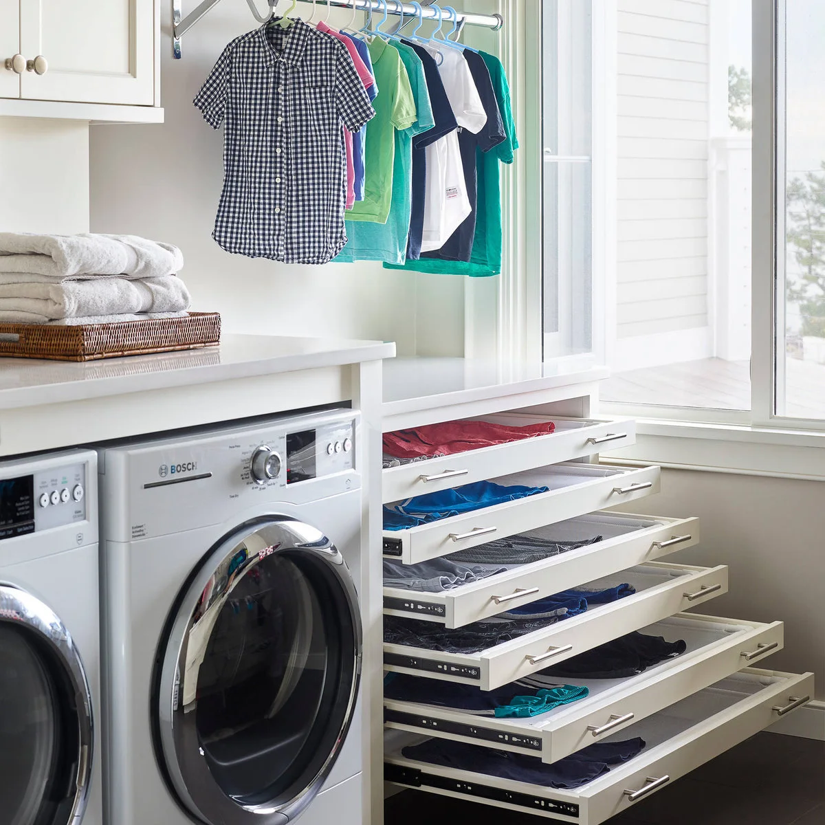 Pull-out mesh drying trays holding clothes inside a white cabinet beside laundry appliances