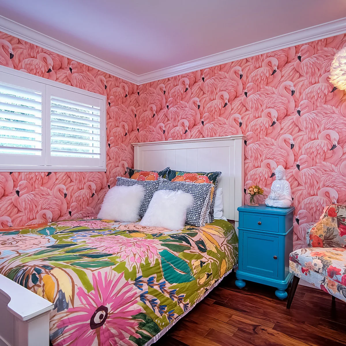 Pink flamingo wallpaper behind a white bed with mixed patterned bedding and a turquoise cabinet