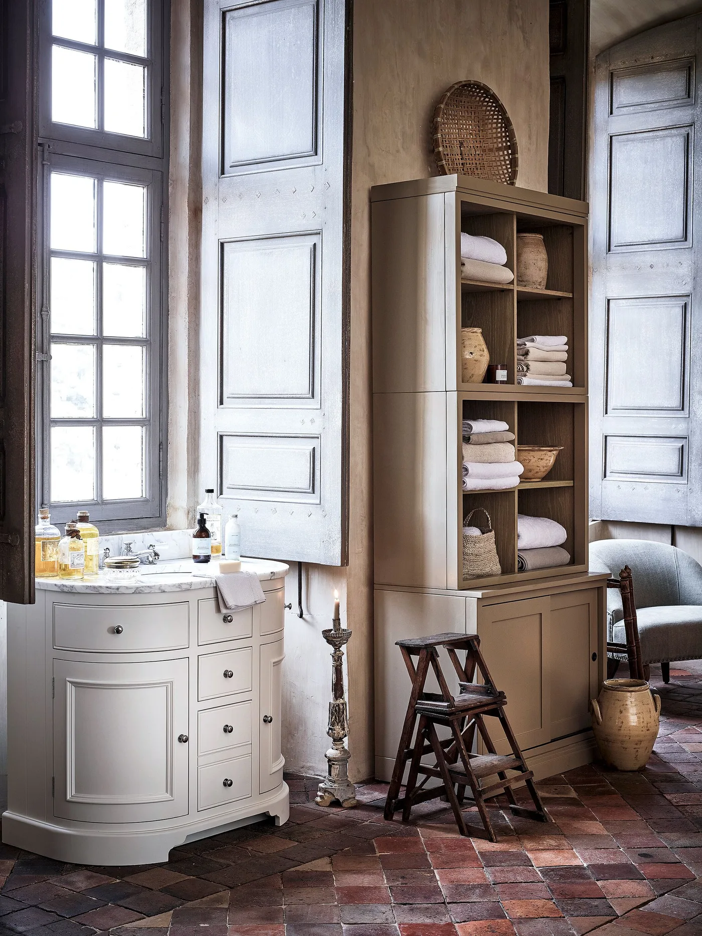 Bathroom with a curved pale vanity, tall open towel cabinet, paneled shutters and warm-toned tiled floor.