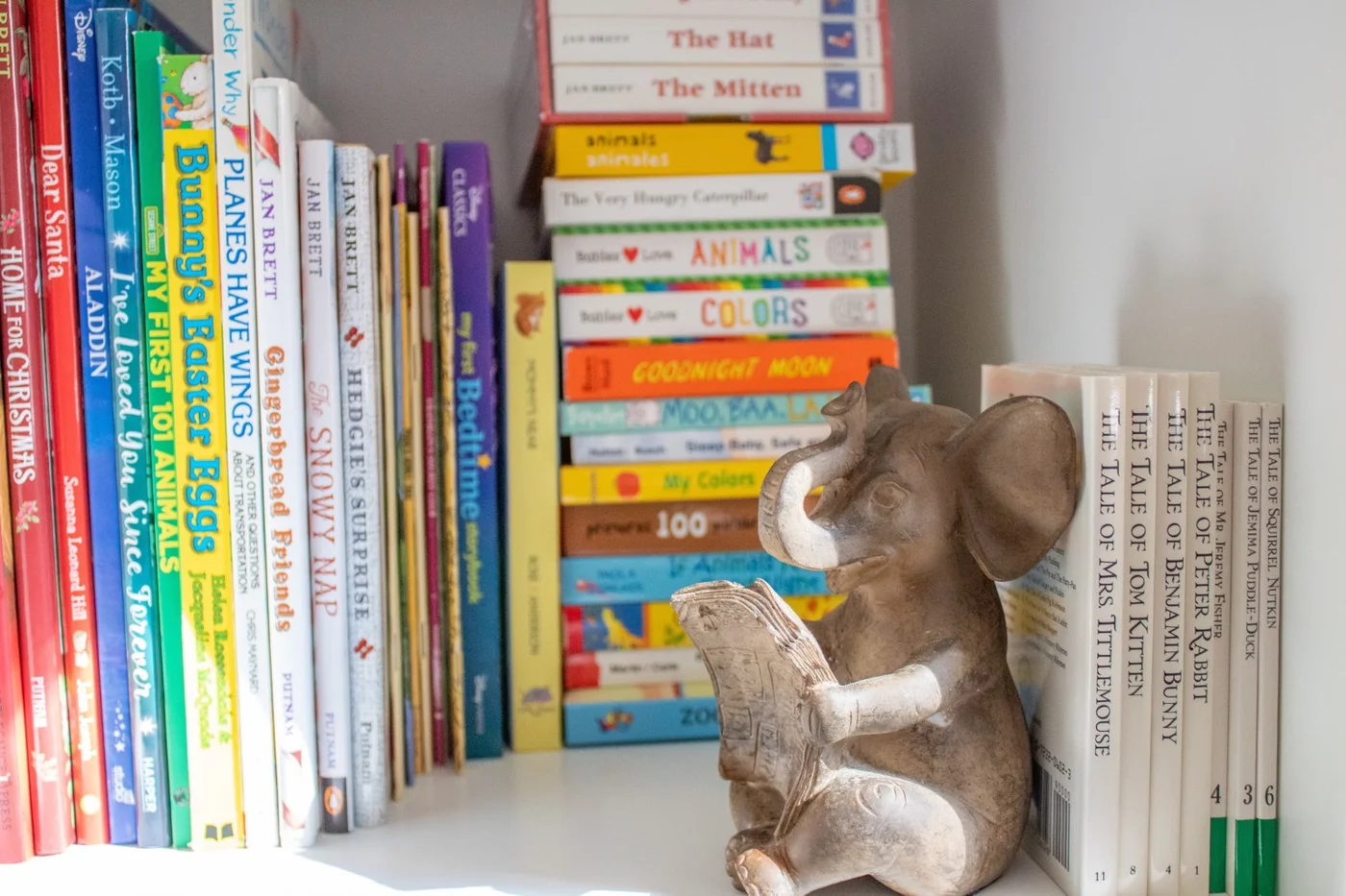 A small elephant figure holding a book sits among upright and stacked children’s books on a white shelf.