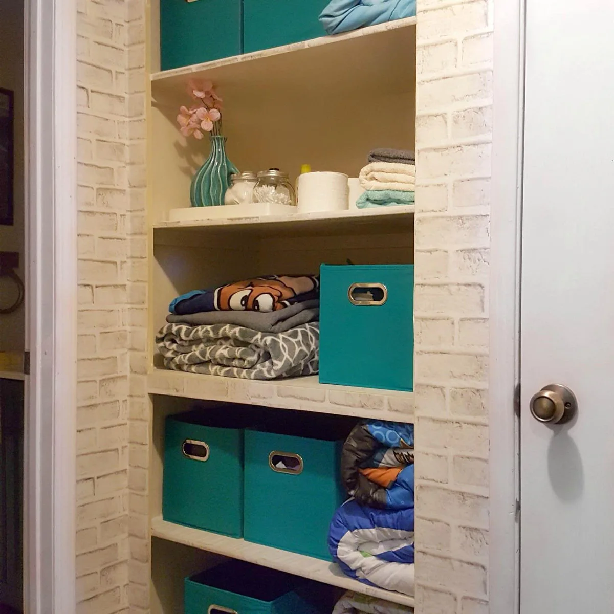 Linen cupboard with turquoise fabric bins, folded towels and patterned bedding against pale brick-pattern walls