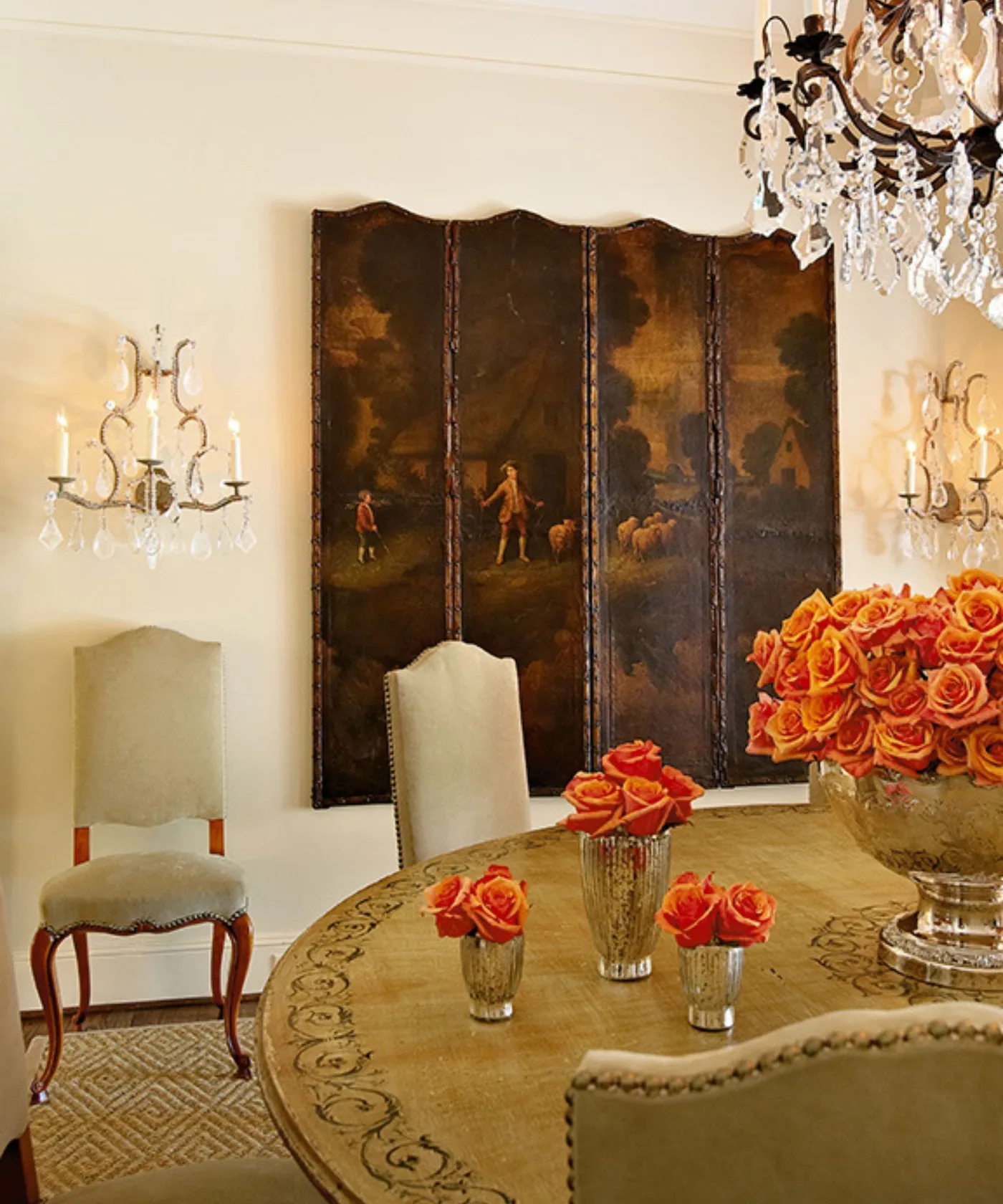 Decorative round dining tabletop with pale curved chairs, a painted folding screen, wall lights and orange roses.