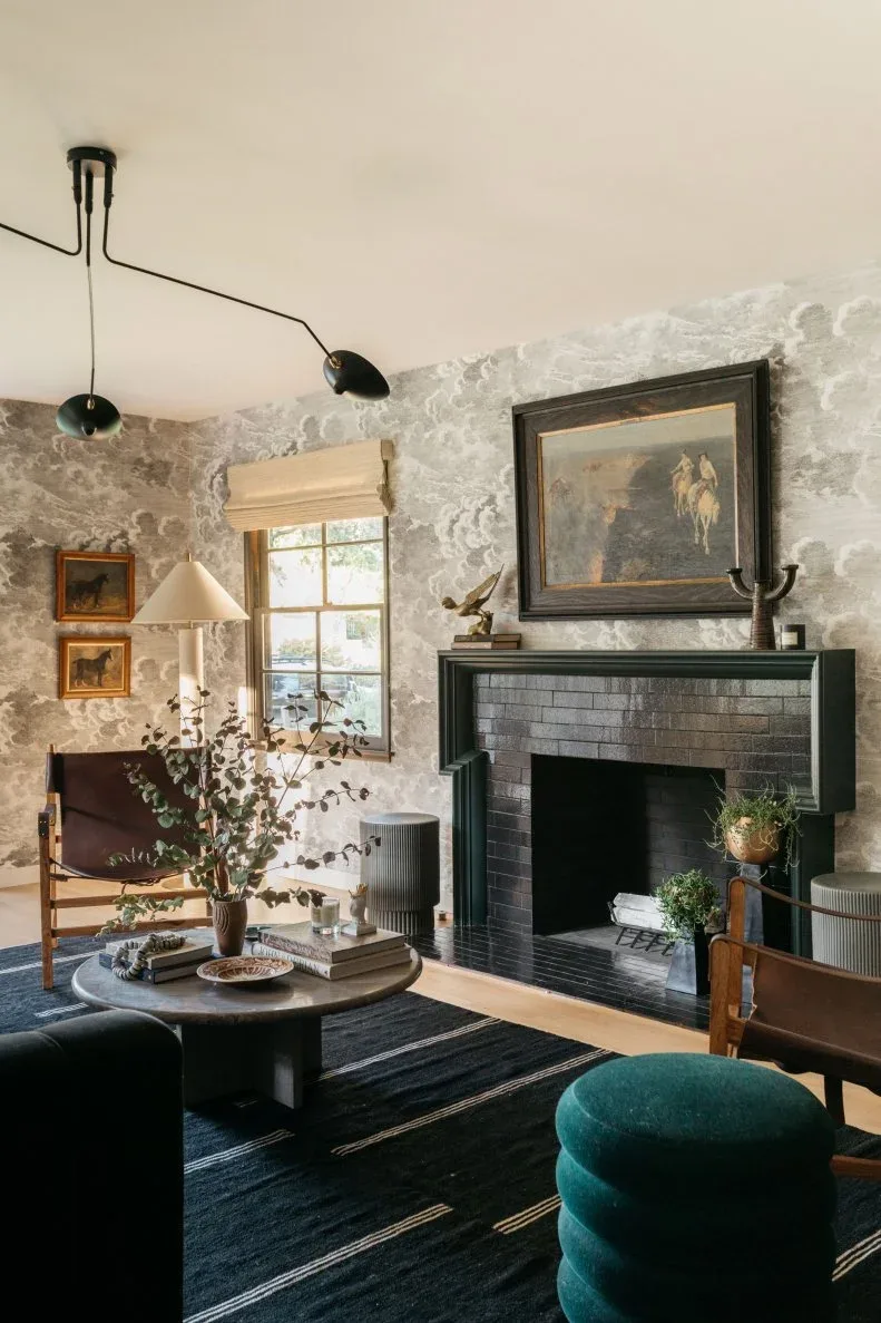 Dark brick fireplace with a large framed painting, pale patterned wallpaper and a green round ottoman.