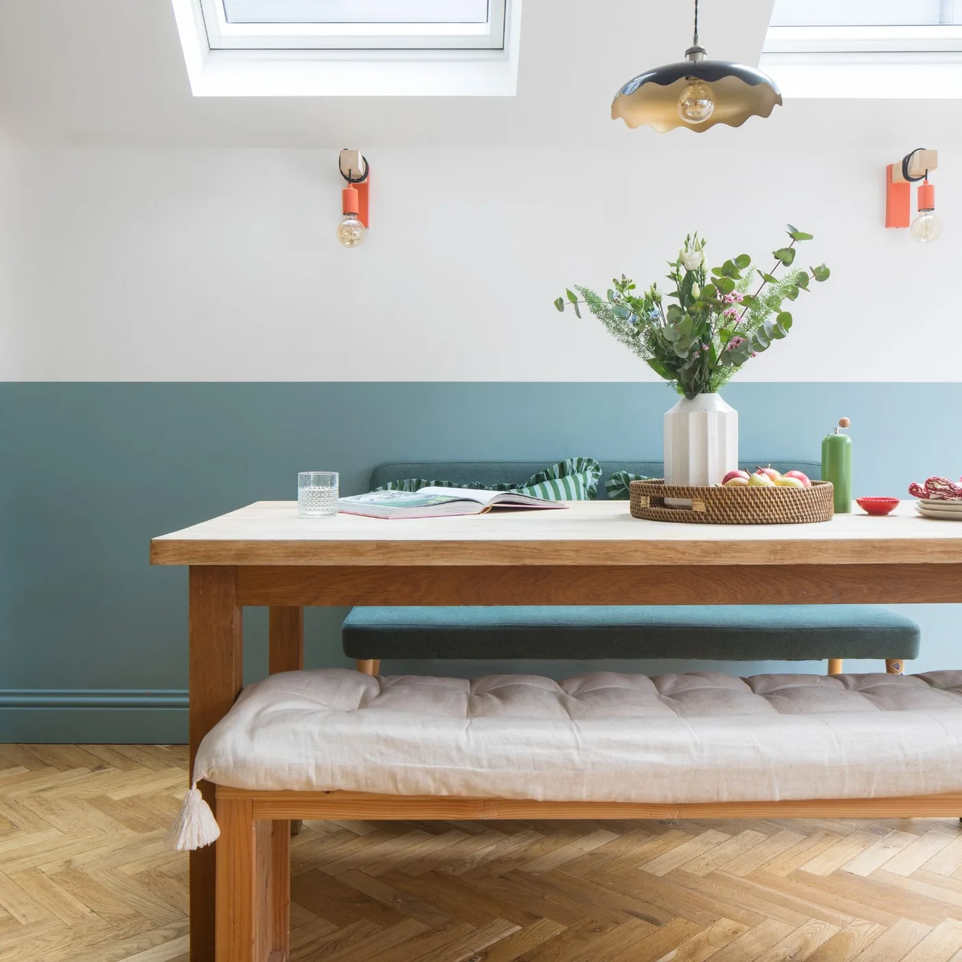 Dining room with pale wood furniture, upholstered benches, blue and white walls, roof windows and a scalloped dark pendant.