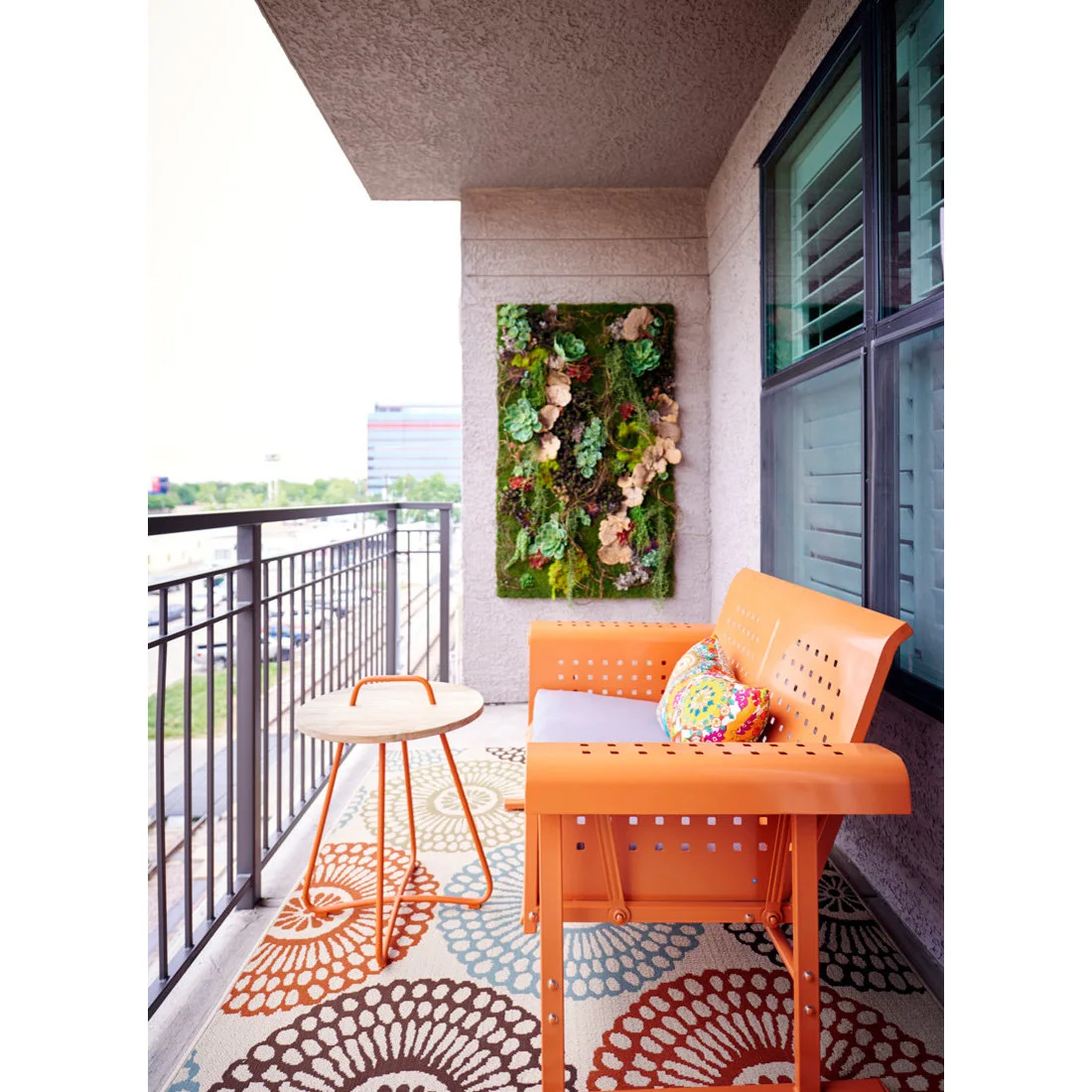 An orange bench and a slim round table sit on a narrow balcony with a floral rug
