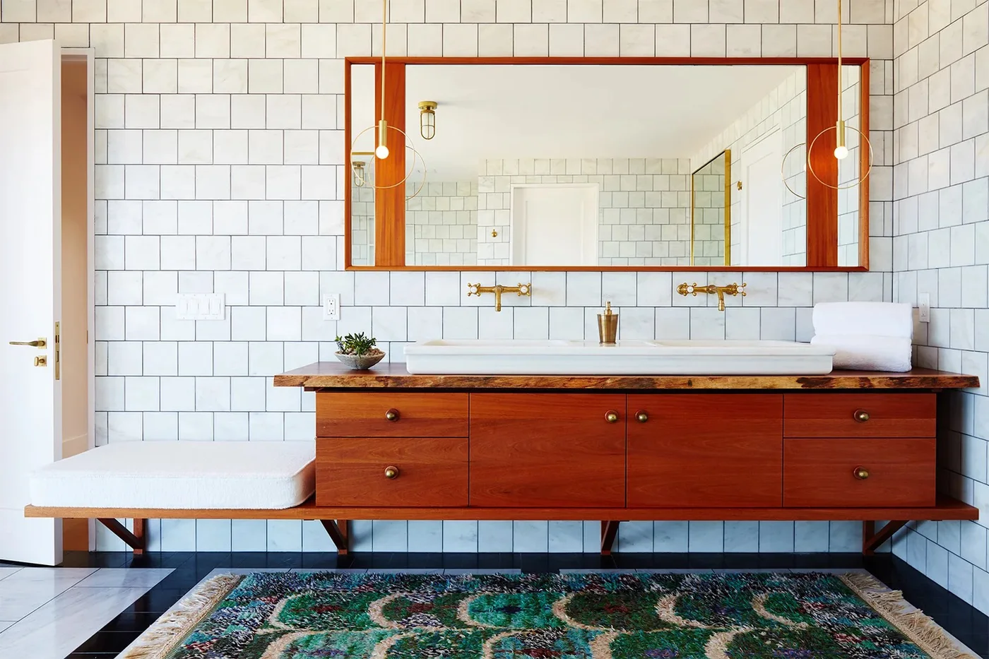 A low wood bathroom vanity supports a long white basin beneath round pendant lights beside a wood-framed mirror and white tiled walls