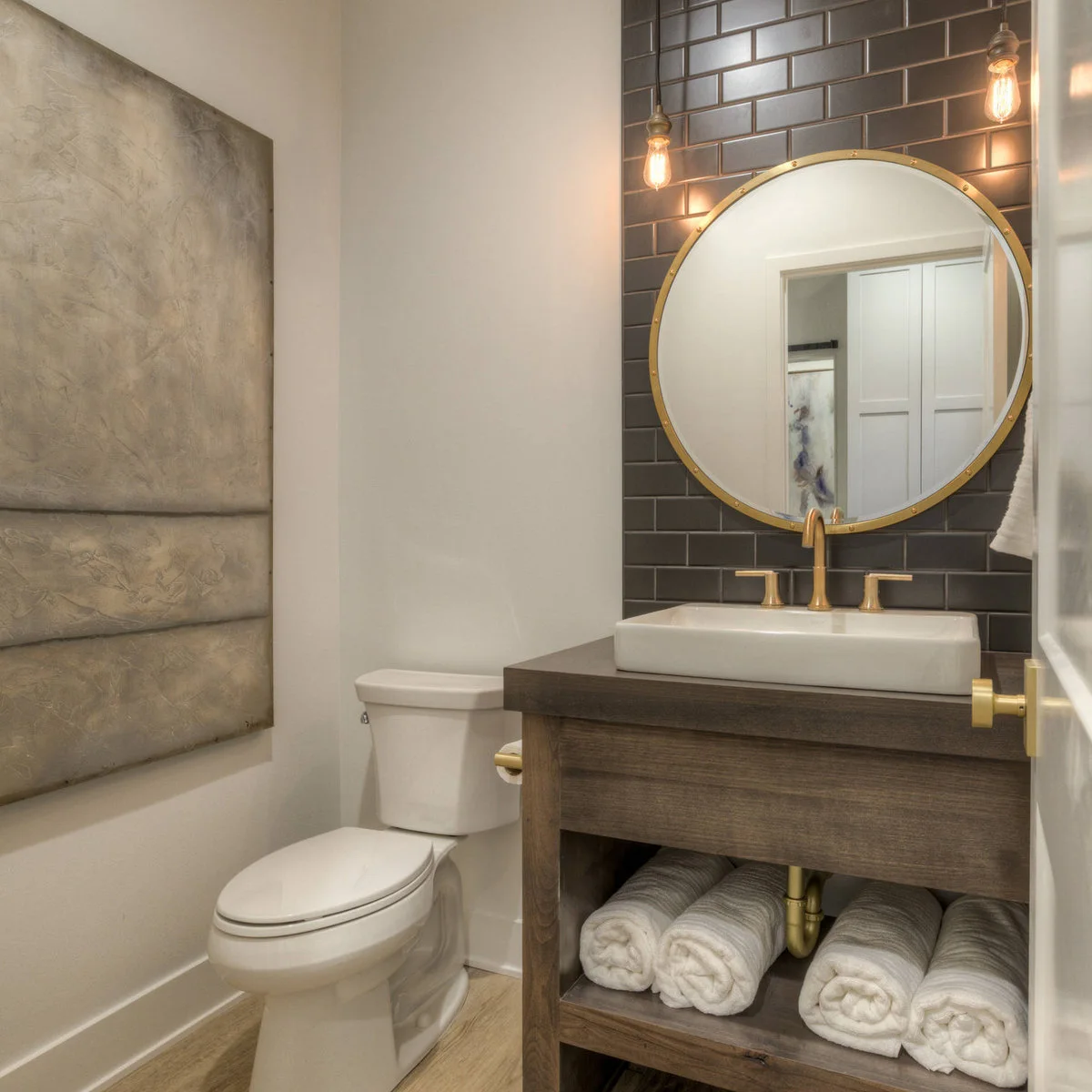 A round mirror and white vessel basin against dark rectangular tile, with a wood console and rolled towels below.