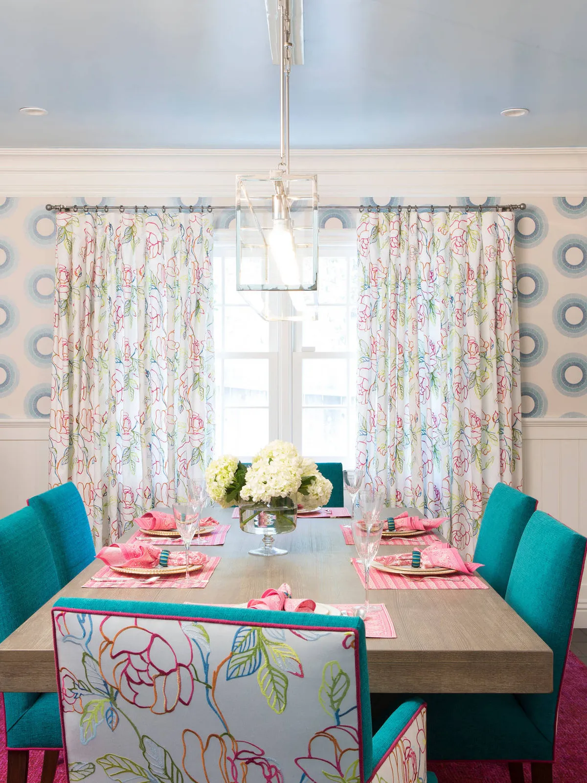 Soft patterned walls and curtains surround bright teal dining chairs with pink accents.