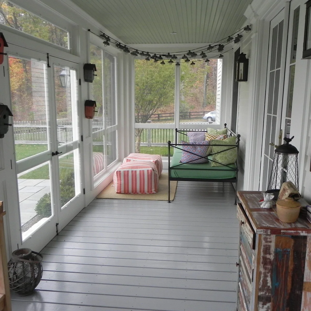 A narrow enclosed porch with gray floorboards, a green-cushioned bench, striped ottomans and overhead string lights.