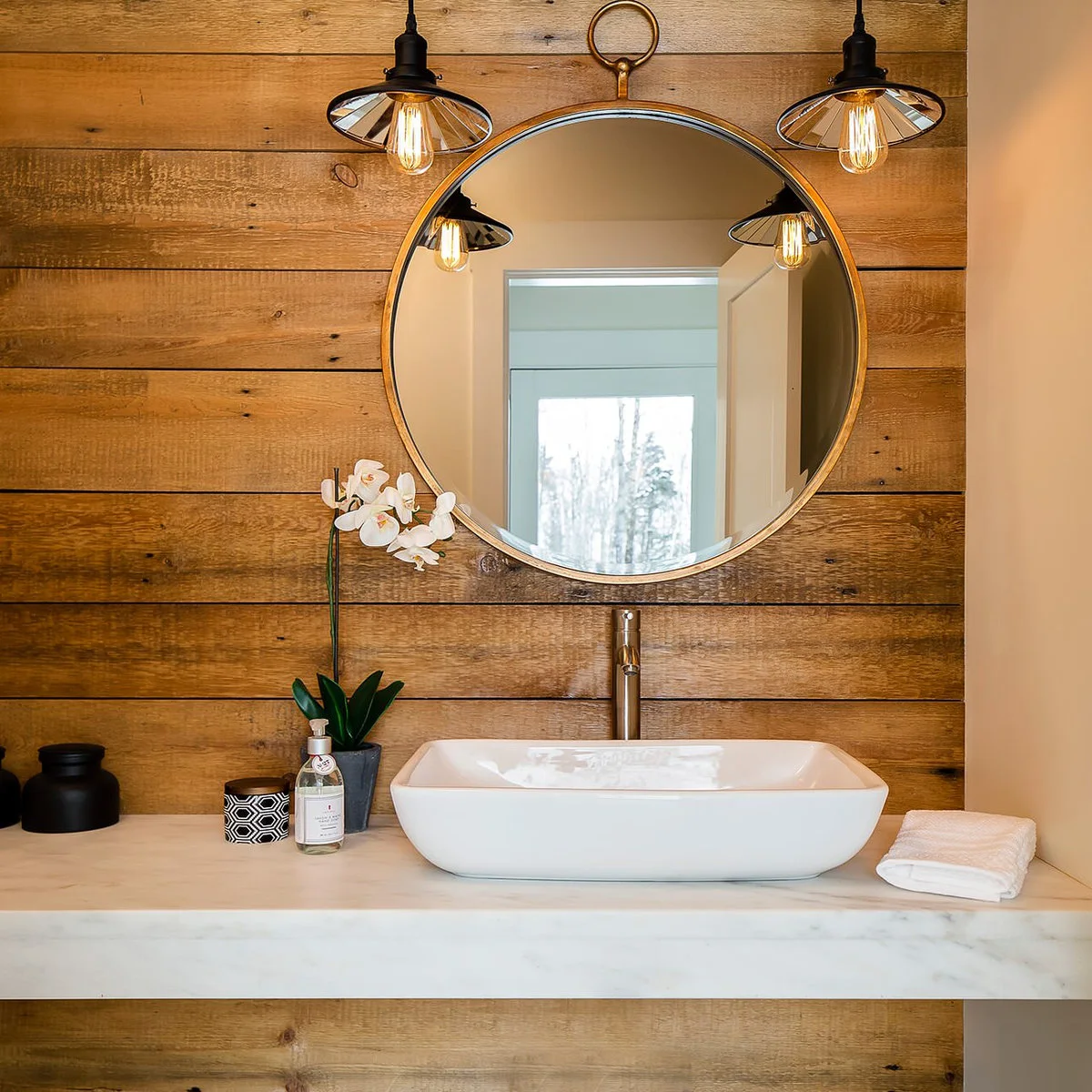 A low white rectangular vessel sink sits on a pale counter below a round metal-framed mirror against warm horizontal wood boards.