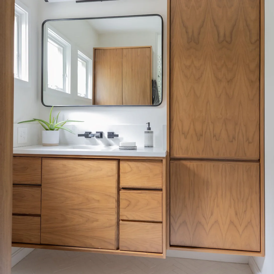 A wood vanity with drawers sits beside a matching tall cupboard, beneath a wide black-edged mirror and a small high window.