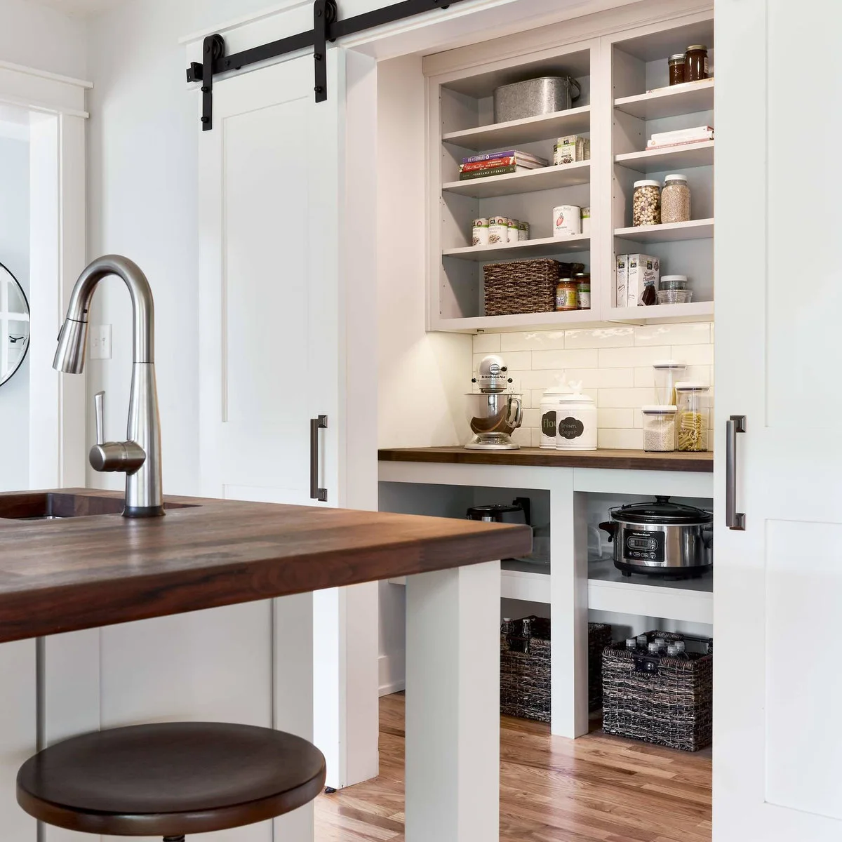 Two white sliding panels open onto a pantry counter with jars and a mixer, with large baskets and stored appliances below.