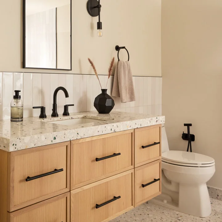 A light wood vanity with black handles supports a speckled counter beneath a slim black mirror beside pale walls.
